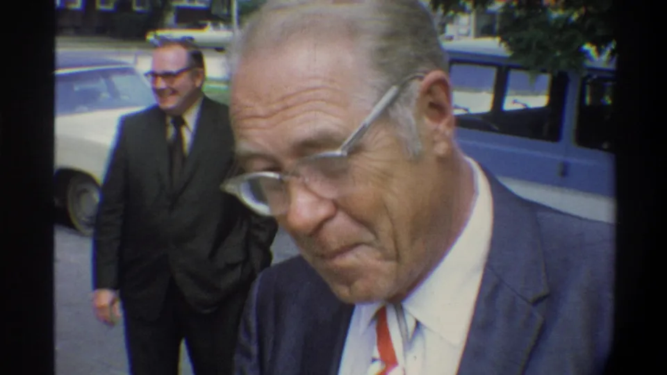 Older man in blue suit, white shirt, and red tie with wire-frame glasses in foreground close-up; second suited man and cream sedan out of focus behind him, Gainesville Florida area, circa 1974.