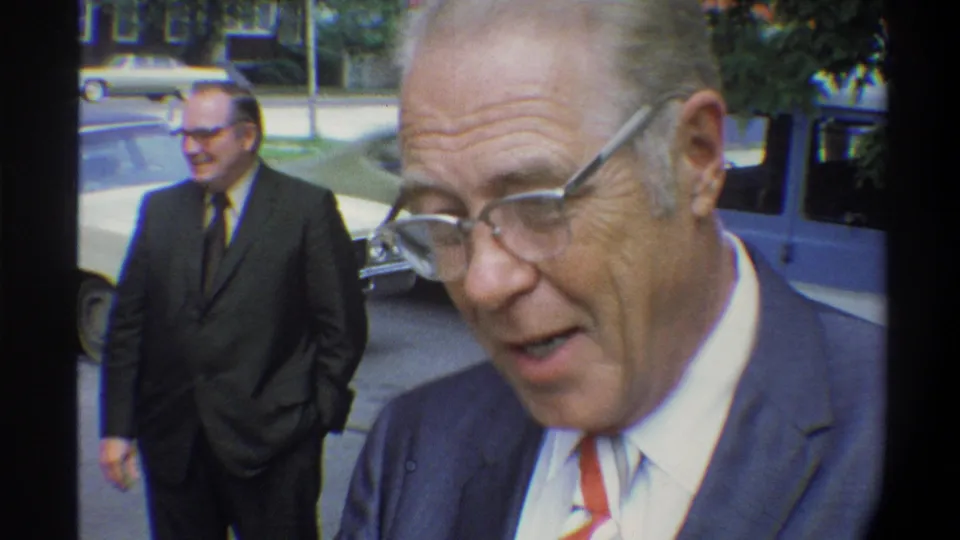 Older man in blue suit, white shirt, and red-and-white tie speaks to camera outdoors with mouth open; second man and cream sedan visible behind him, Gainesville Florida area, circa 1974.