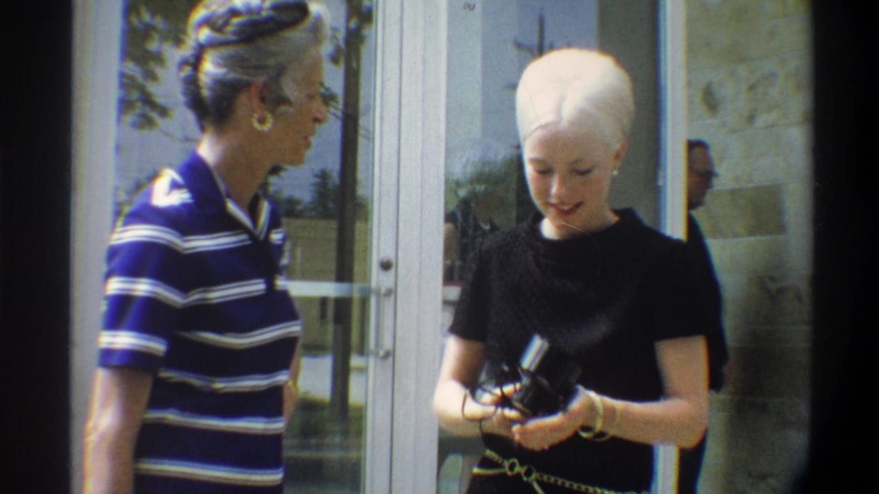 Two women in mid-1970s dress converse outside a stone and glass-door building entrance, likely Gainesville, Florida, circa 1974; one holds a compact handheld camera.