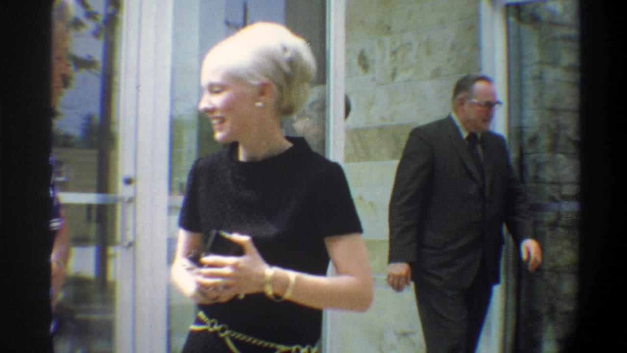 Platinum-blonde woman in black top laughs and gestures outdoors; a man in a dark suit passes behind her near glass doors.