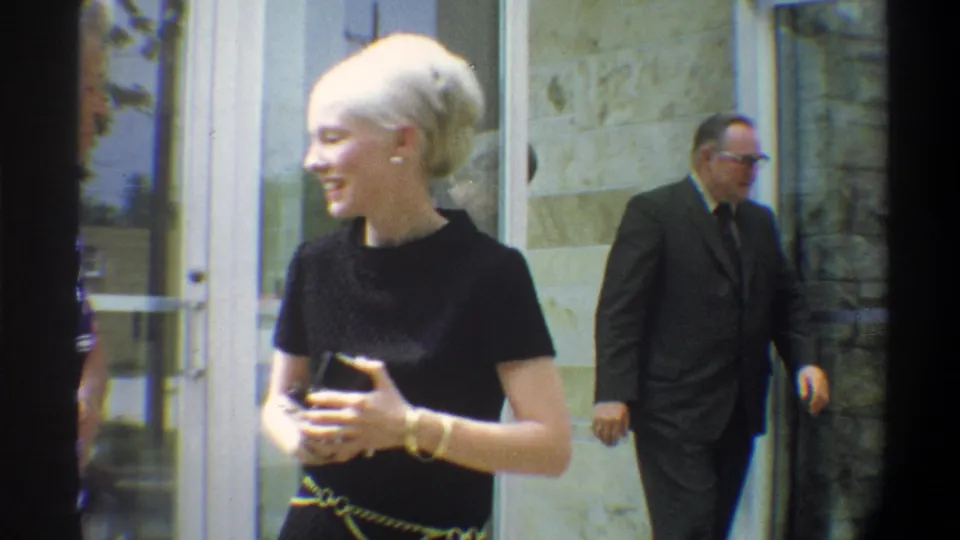 A platinum-blonde woman in a black top with gold chain belt laughs and gestures outside a stone building, circa 1974 Gainesville, Florida; a man in a dark suit walks past in the background.
