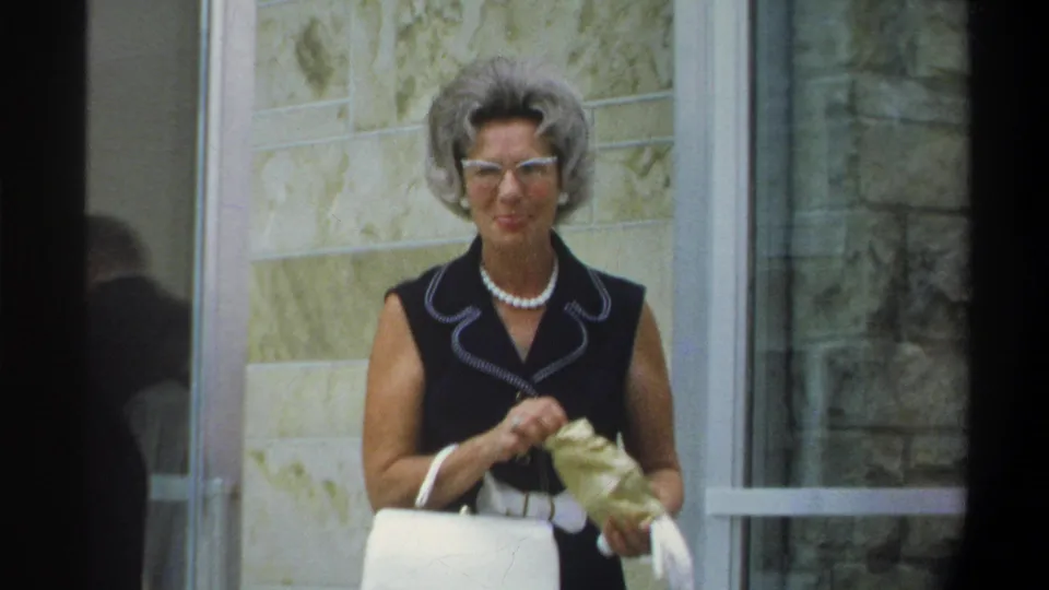 An older woman in a sleeveless black top with white piping trim, pearl necklace, and cat-eye glasses stands outside a building entrance, circa 1974 Gainesville, Florida, holding a white handbag.