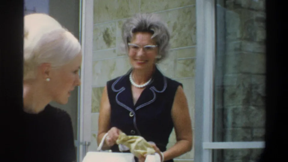Continued view of an older woman with gray hair, cat-eye glasses, and pearl necklace outside the stone building entrance, circa 1974 Gainesville, Florida; another woman's head partially visible at frame left.