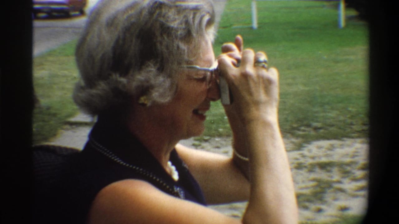 Older woman with styled gray hair, blue-framed cat-eye glasses, and dark navy dress with beaded trim raises a compact camera while laughing; suburban outdoor setting, mid-1970s.