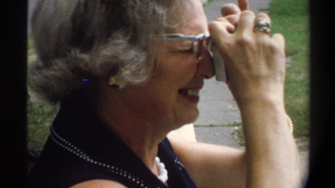 Close-up of older woman in cat-eye glasses holding a compact camera to her eye, ring visible on her raised hand.
