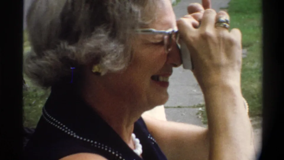 Close-up of older woman with blue-framed glasses and styled gray hair holding camera to eye, ring on raised hand; mid-1970s 8mm film, suburban outdoor setting.