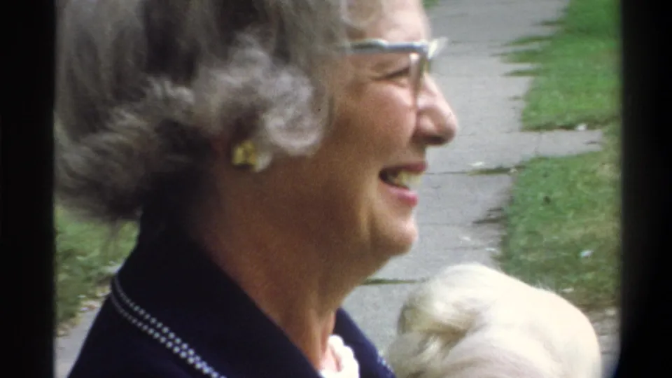 Older woman with styled gray hair and cat-eye glasses laughing heartily outdoors with camera lowered; a second lighter-haired figure is partially visible at bottom of frame, mid-1970s.