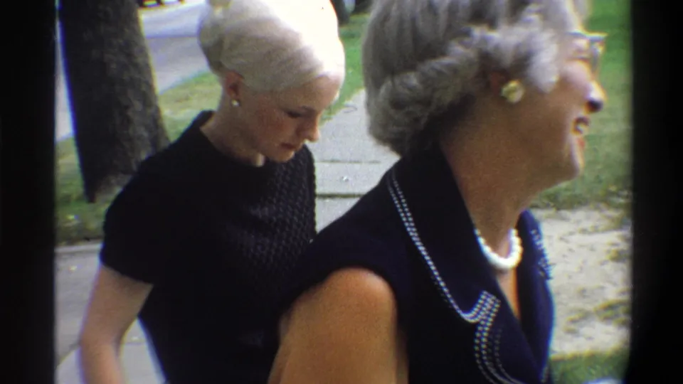 Two women on a suburban sidewalk: gray-haired older woman in dark navy dress with beaded trim laughing at right, second lighter-haired woman bending forward at left; Gainesville, Florida, 1974.