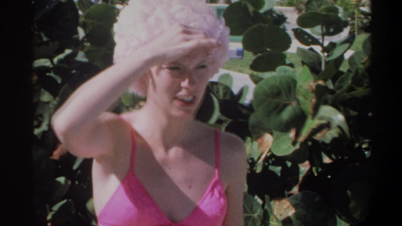 Young woman in pink string bikini and ruffled swim cap shielding eyes from bright sunlight, lush tropical foliage behind her.