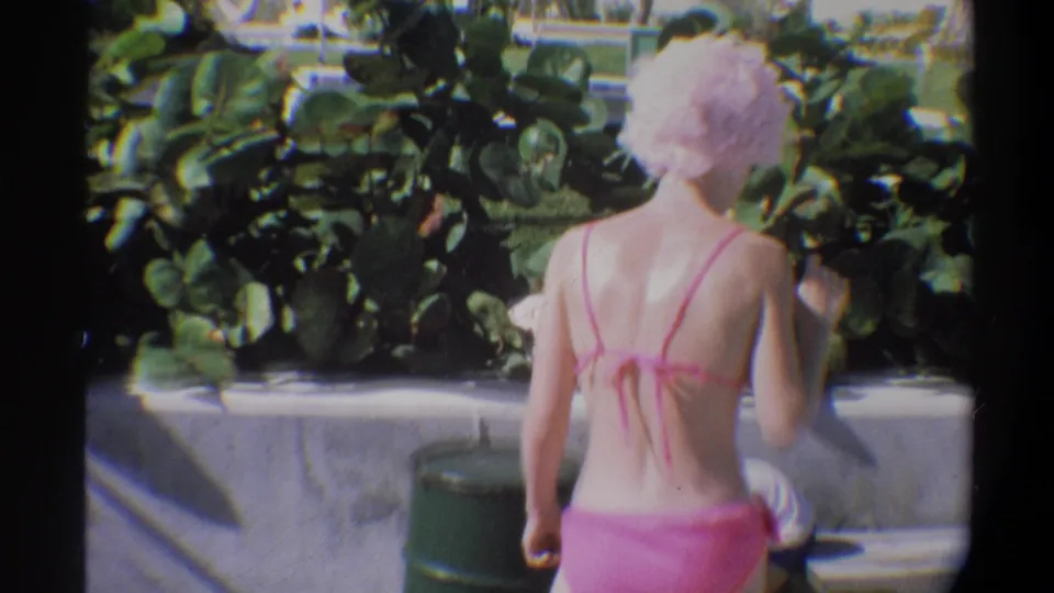 Young woman in pink string bikini walking away toward a pool, concrete pool surround and dense tropical shrubs visible, Gainesville Florida area, circa 1974.