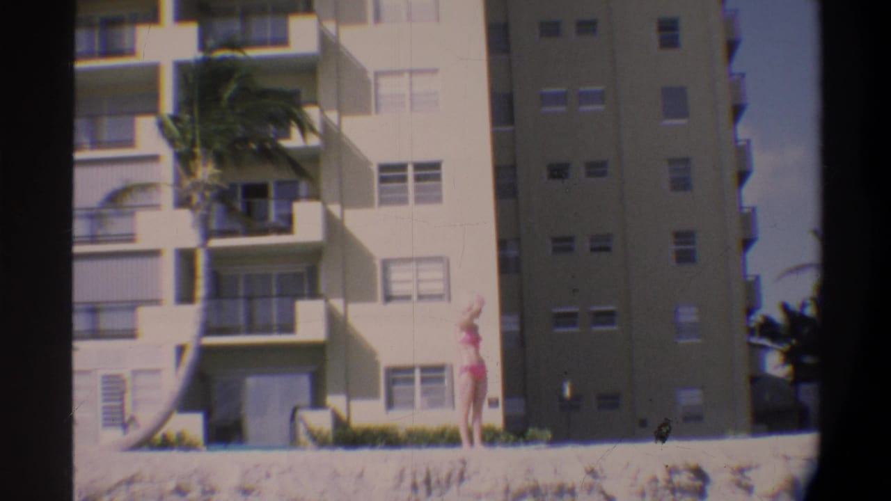Woman in pink bikini and matching hat stands near concrete wall; multi-story apartment building and palm tree behind her, 8mm vignette.