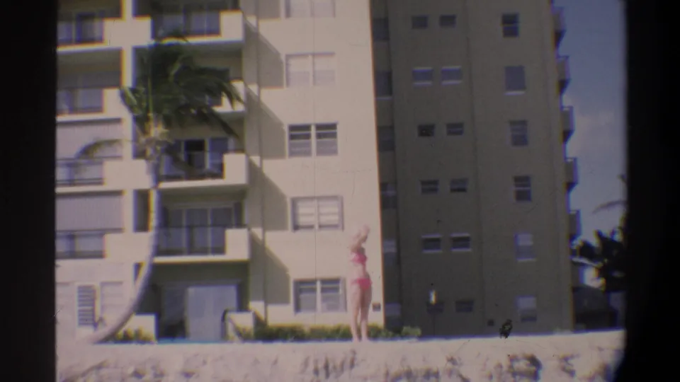 Woman in pink bikini and wide-brim hat poses near a concrete ledge with a Florida mid-rise apartment building and palm tree in the background, circa 1974.