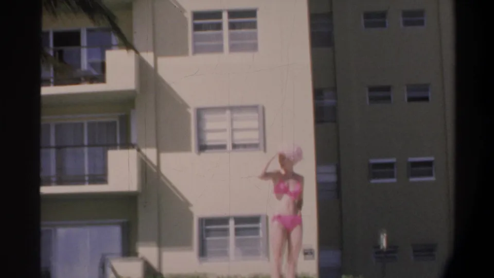 Woman in pink bikini adjusts her wide-brim hat while standing before a multi-story Florida apartment building with balconies, circa 1974.