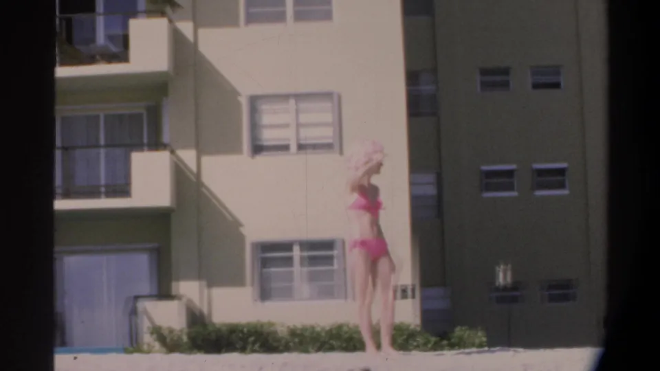 Woman in pink bikini poses near a low concrete wall with a multi-story Florida beach apartment building and palm tree behind her, circa 1974.