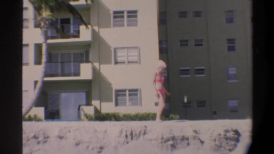 Woman in pink bikini and hat stands by a low concrete structure in front of a Florida beachfront apartment building, circa 1974.