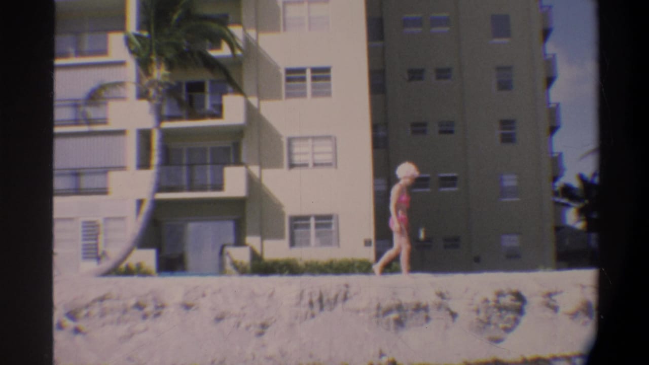 Woman in pink bikini and swim cap walking along a sandy beach in front of a large multi-story building, palm tree at left.