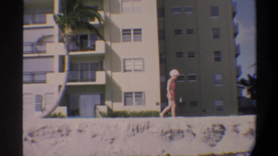 Woman in pink bikini and swim cap strolling along a Florida beach, c. 1974, with a multi-story beige resort building and palm tree behind her.