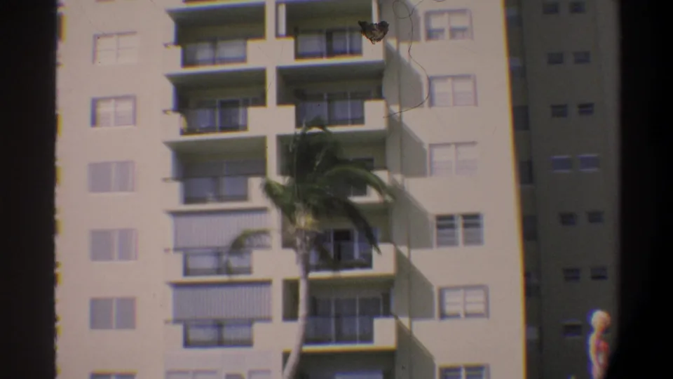 Beachfront high-rise with repeating balcony grid dominates frame; woman in pink bikini visible at right edge, c. 1974 Florida.