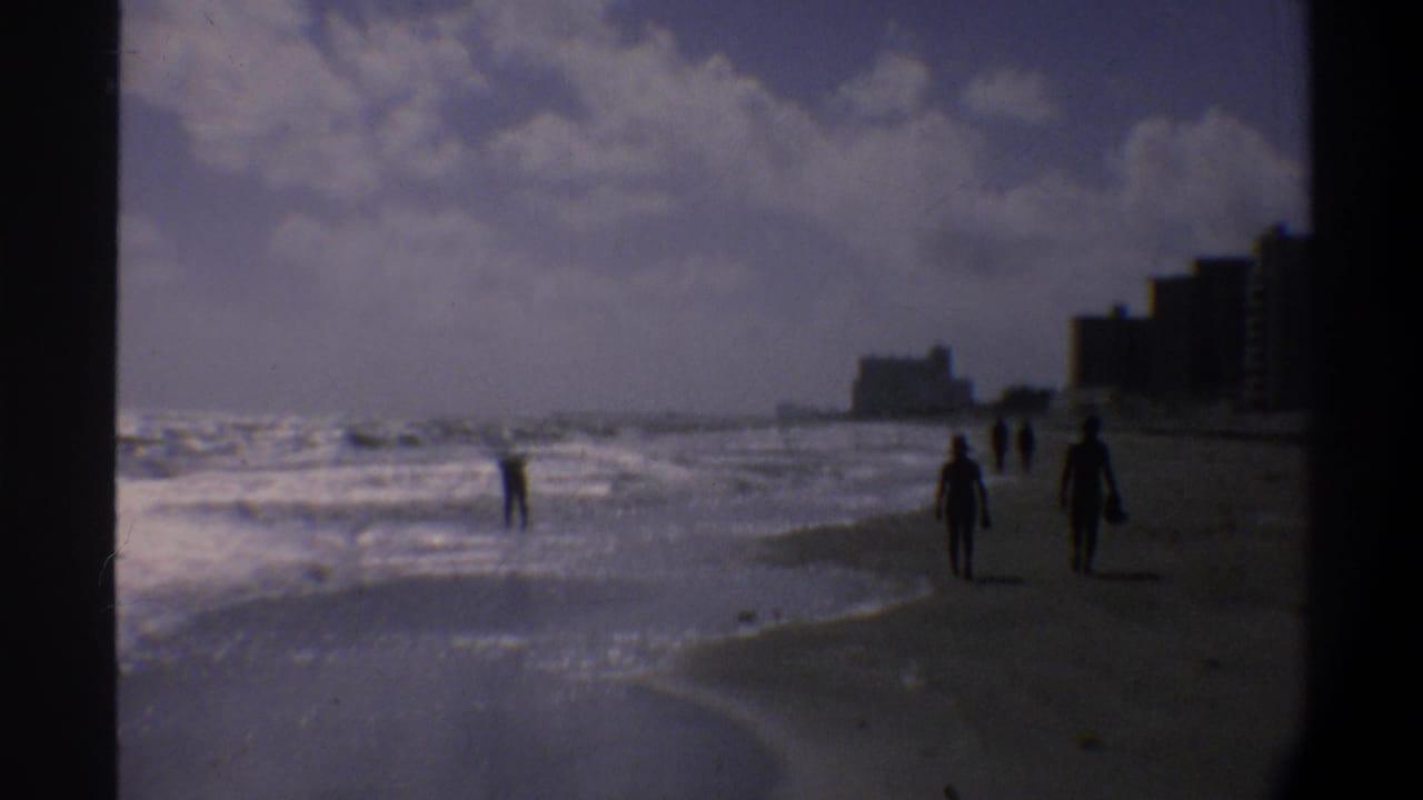 Several silhouetted figures near the waterline on an East Coast Florida beach, circa 1974, with coastal high-rise buildings in the background.