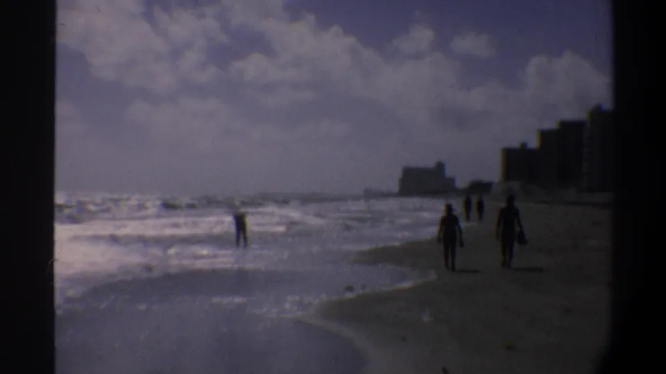 Several silhouetted figures near the waterline on an East Coast Florida beach, circa 1974, with coastal high-rise buildings in the background.