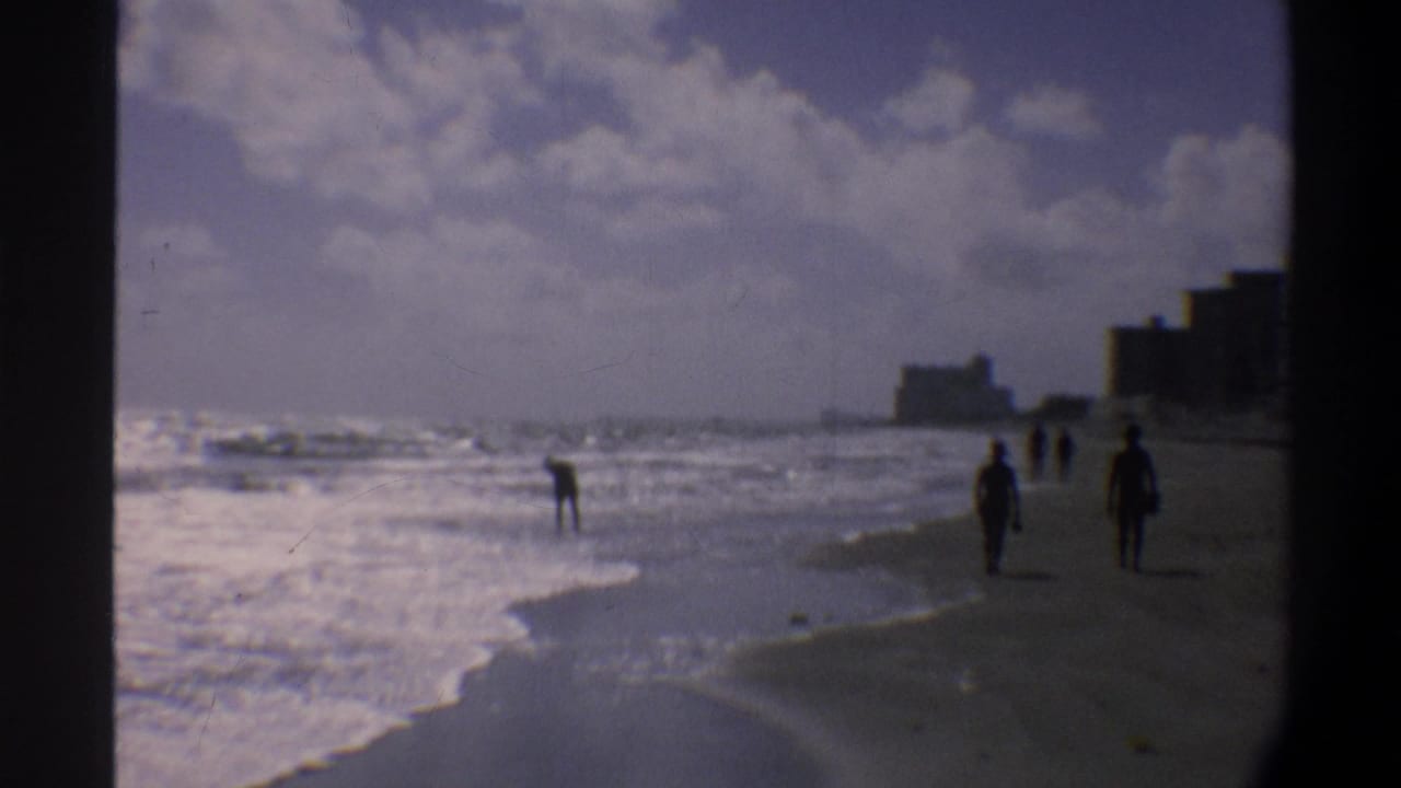 Three silhouetted figures stand near the waterline, with breaking surf and a coastal building skyline behind them.