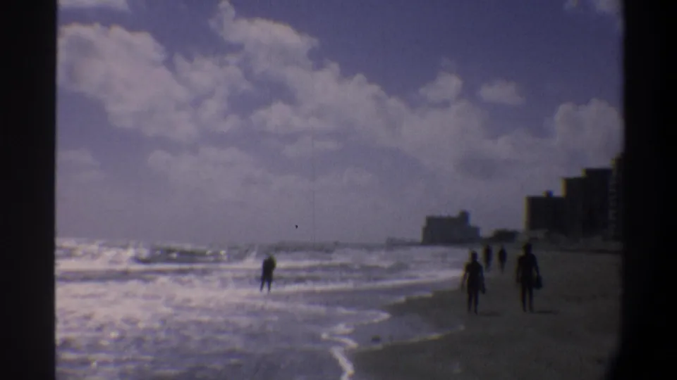 Wide shot of an East Coast Florida beach, circa 1974, showing multiple beachgoers, breaking waves, and a coastal high-rise skyline under large cumulus clouds.