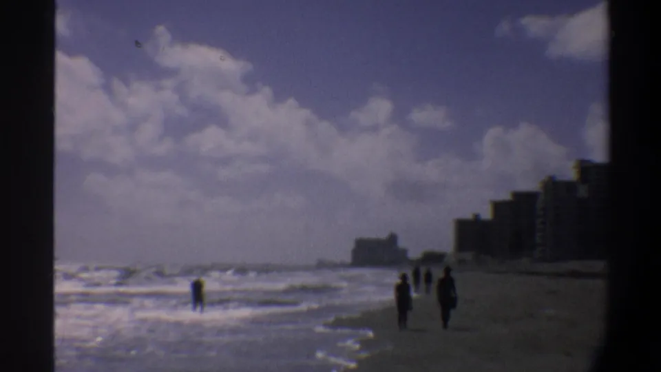 Multiple beachgoers on an East Coast Florida beach, circa 1974, with mid-rise coastal condominiums stretching along the shoreline under a cloudy sky.