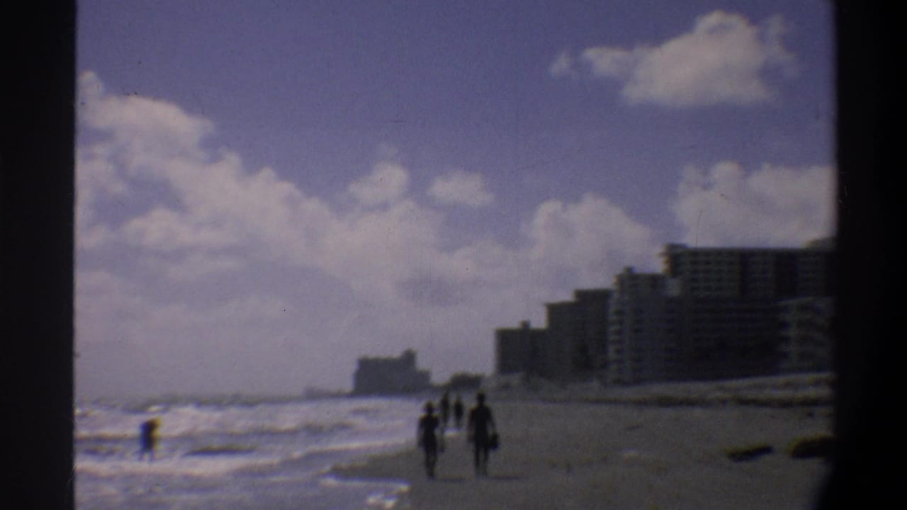 Silhouetted beachgoers stroll near the waterline on a Florida coastal beach, with a row of modernist hotel towers behind them, circa 1974, 8mm film.