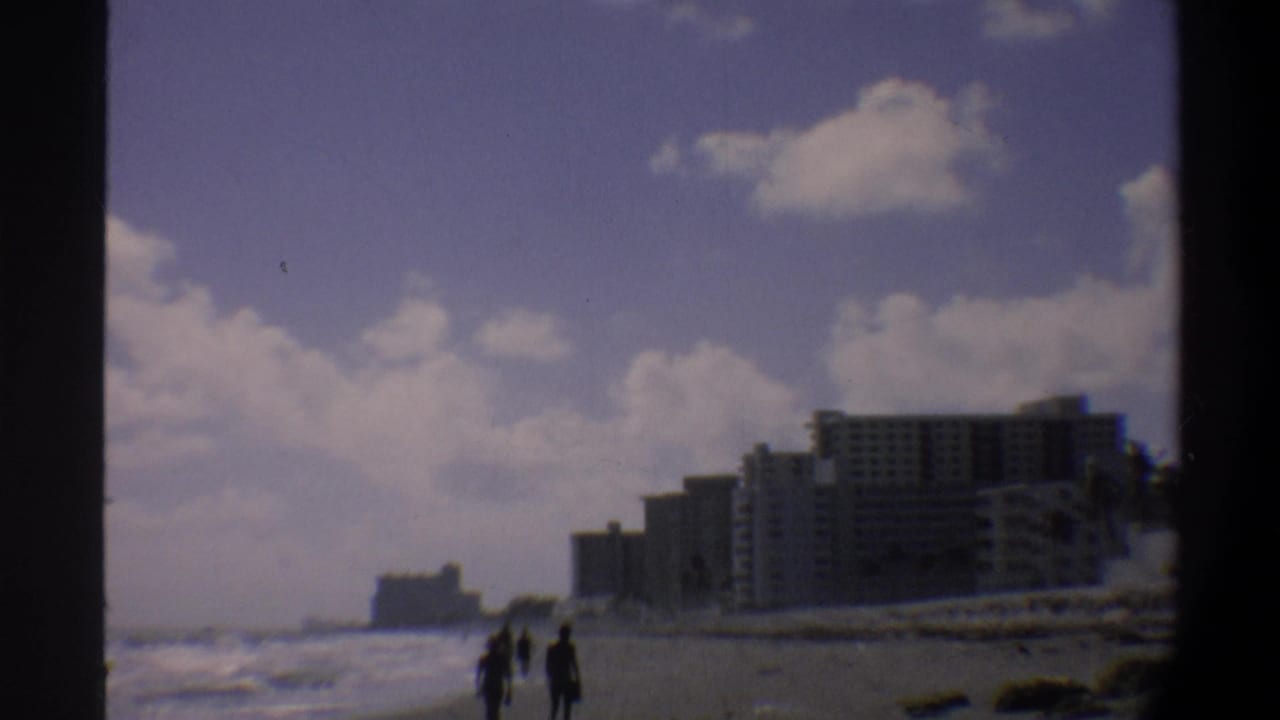 A few pedestrians walk near the waterline on a sandy beach, with blocky mid-rise hotels visible behind them, 8mm color.