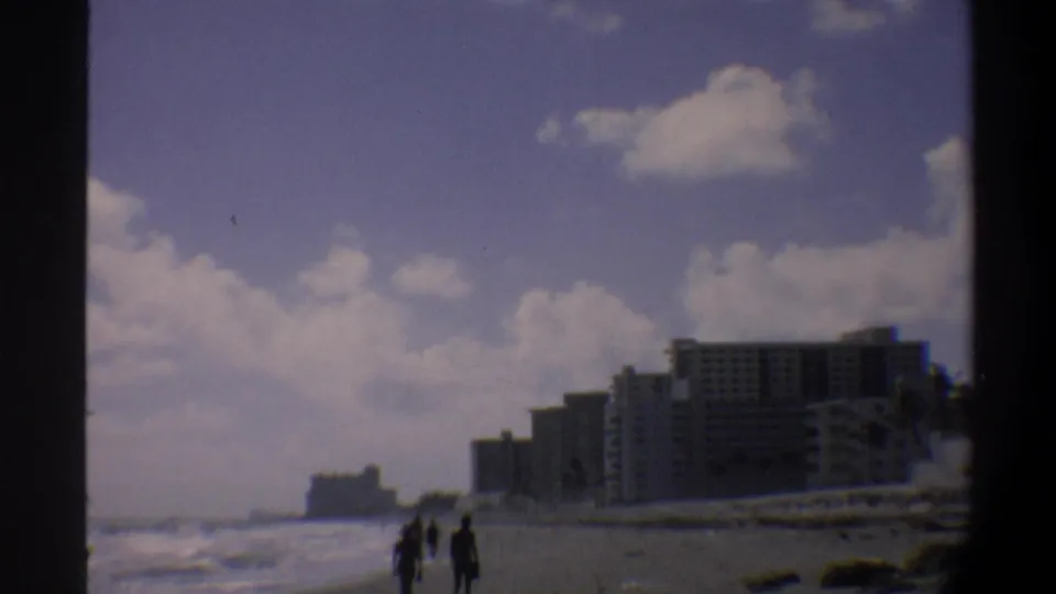 Pedestrians walk along a Florida beach in silhouette with 1970s-era beachfront hotel towers forming the backdrop, circa 1974, 8mm footage.