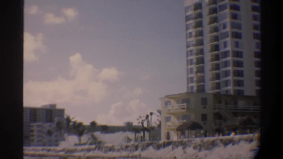 A tall modernist white high-rise hotel with balconies rises above palm trees on a Florida beach, with secondary resort buildings behind, circa 1974, 8mm film.