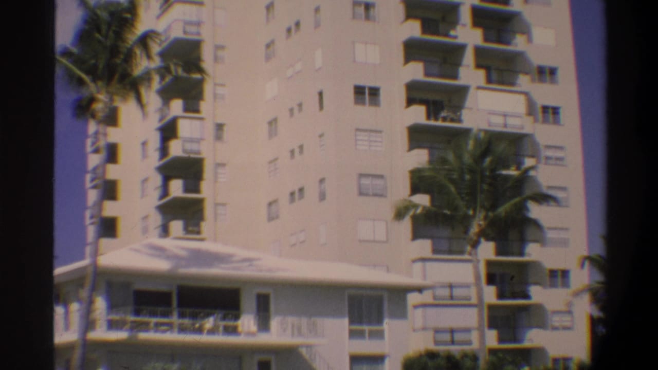 Same high-rise building slightly reframed right, balconies and palm trees visible, lower terrace still in shot, 8mm home movie.