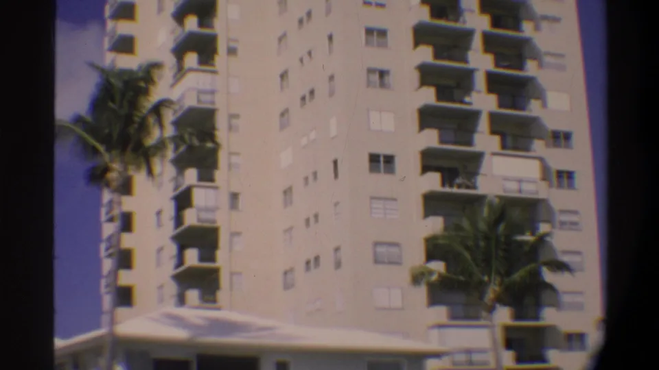 8mm home-movie frame tilting up the facade of a multi-story Florida building; balconies and palm fronds dominate, lower terrace no longer in view, circa 1974.