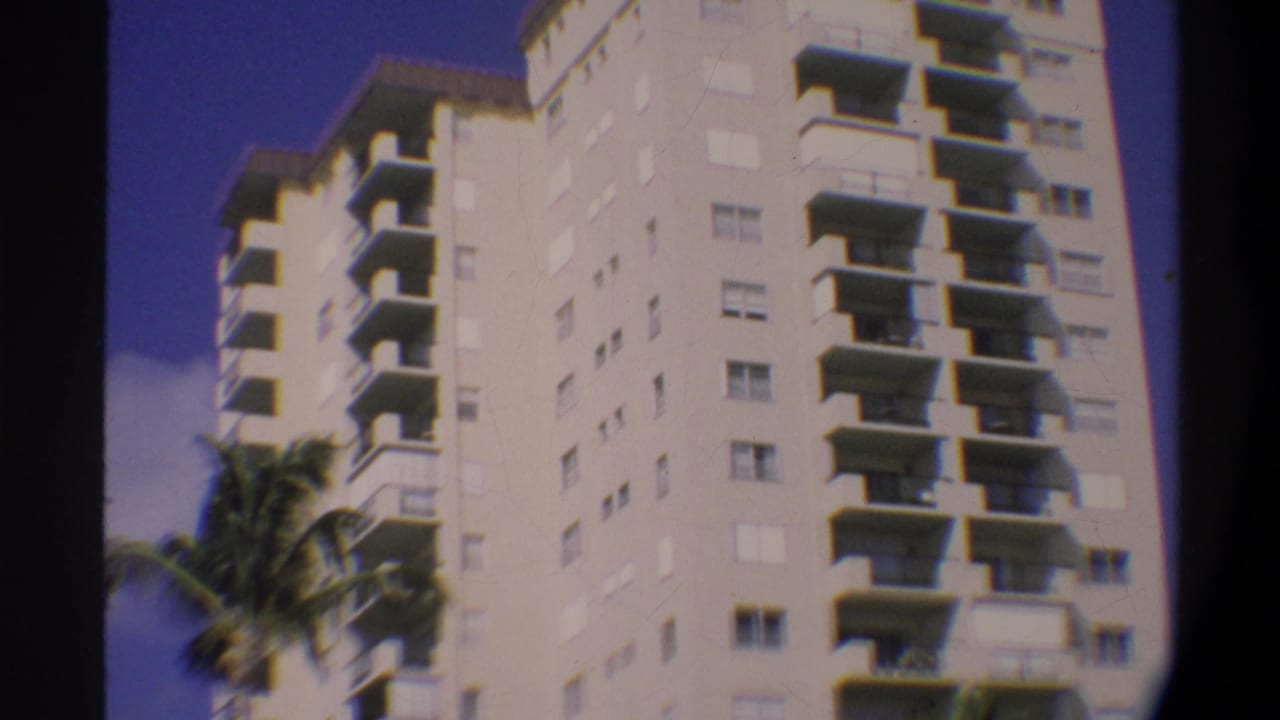 Low-angle 8mm home movie view of a white multi-story Florida high-rise, circa 1974, with palm fronds in the foreground and blue sky above.