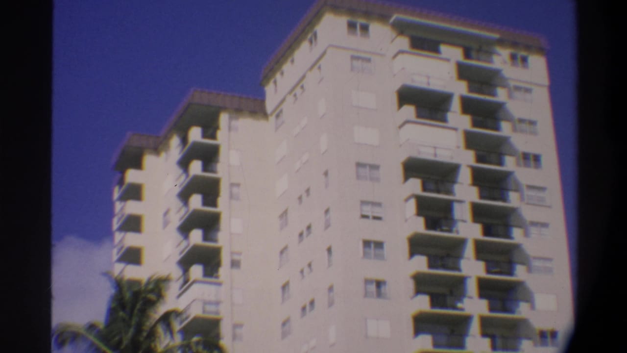 Reframed 8mm view of the same white high-rise with palm fronds at lower right and a partly cloudy blue sky.