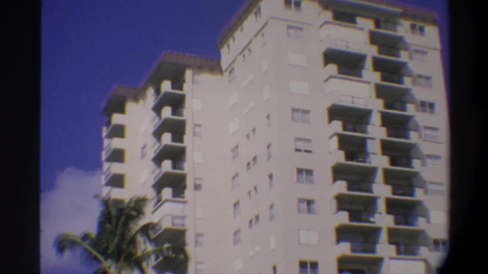 Closer low-angle 8mm home movie view of balcony tiers on a Florida high-rise, circa 1974; palm fronds in foreground and blue sky above.