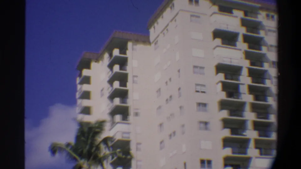 Street-level 8mm view of a white Florida residential high-rise, circa 1974; palm fronds at lower right and a cloud-dotted blue sky in the same continuous shot.