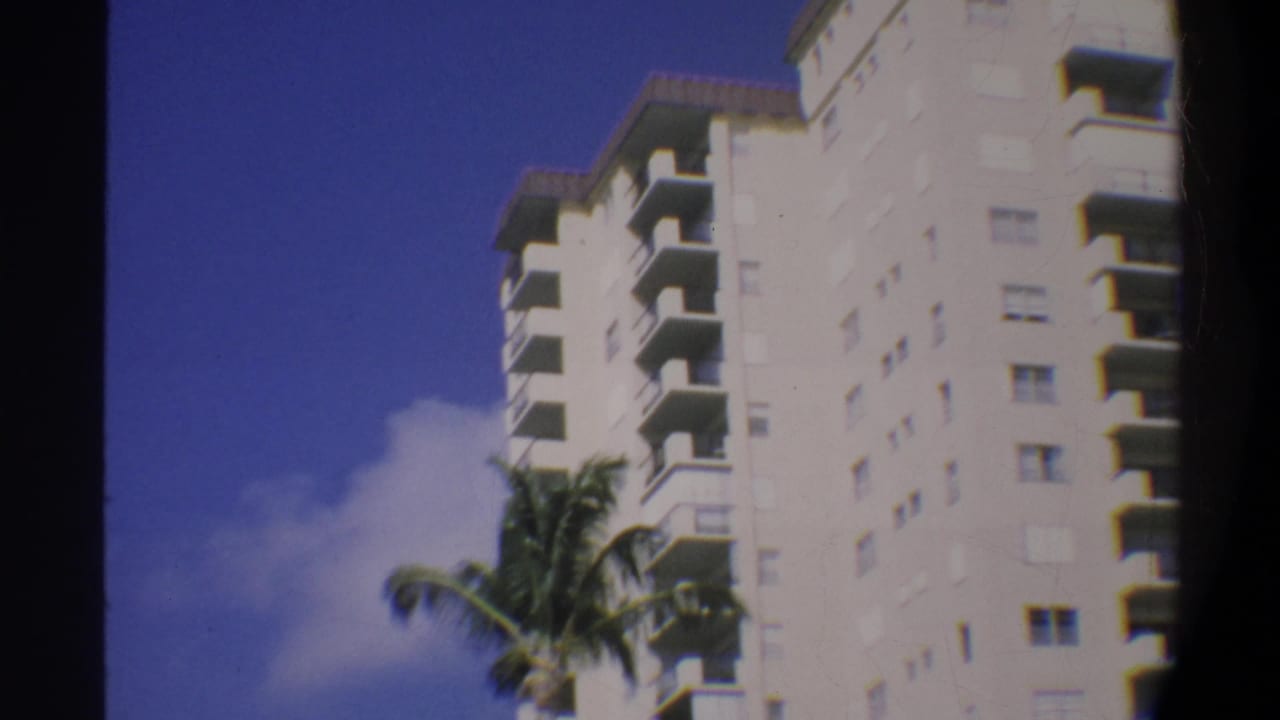 Super 8 home movie frame showing a tall white apartment building with repeating balconies in Gainesville, Florida, 1974; palm fronds and blue sky visible.
