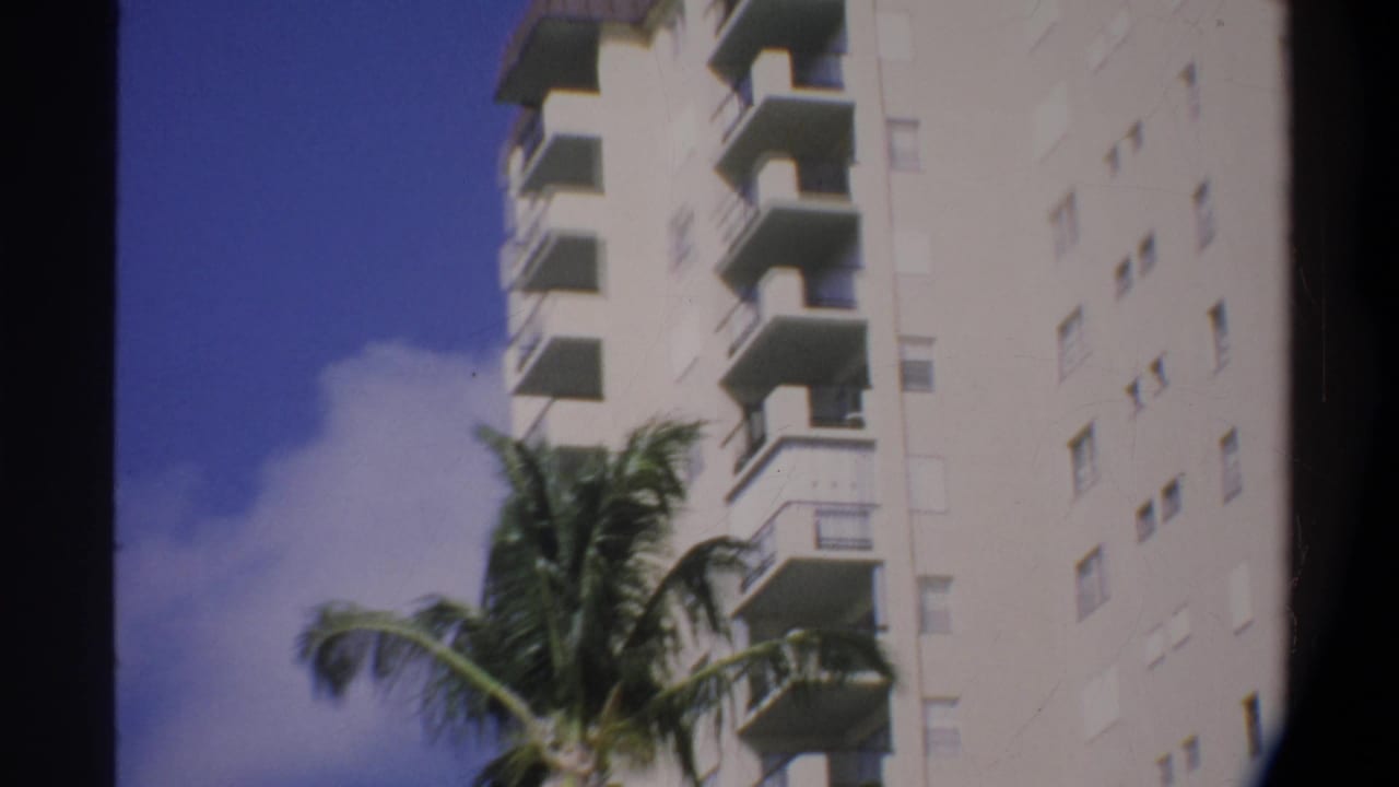 Tall white apartment building with stacked balconies, palm fronds at left, bright blue sky, 8mm home movie grain.