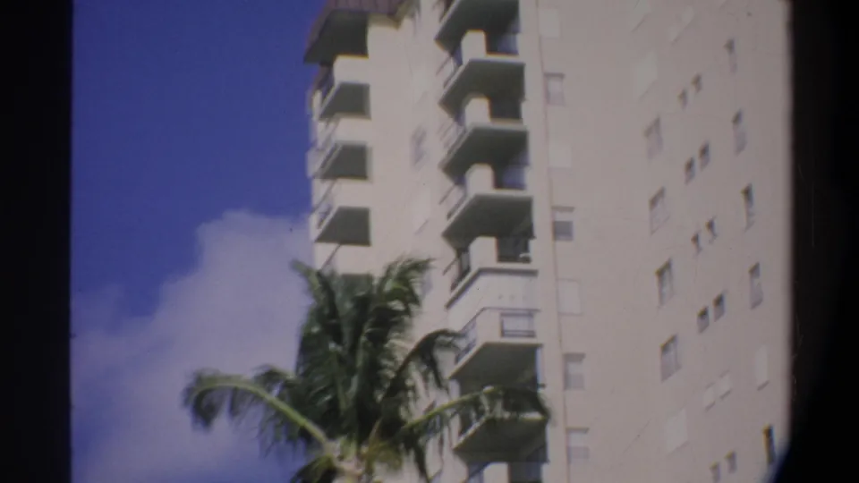 Super 8 frame of a multi-story white apartment building with repeating balconies, palm fronds, and blue sky; consistent with 1974 Florida home movie footage.