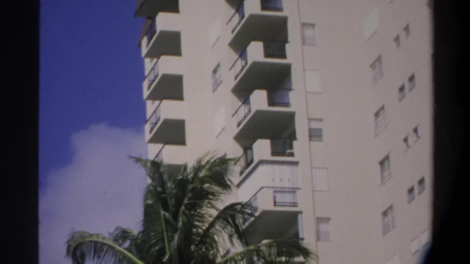 Super 8 home movie frame showing a low-angle view of the apartment building's lower floors with railing details and palm fronds in foreground, Gainesville, Florida, c.1974.