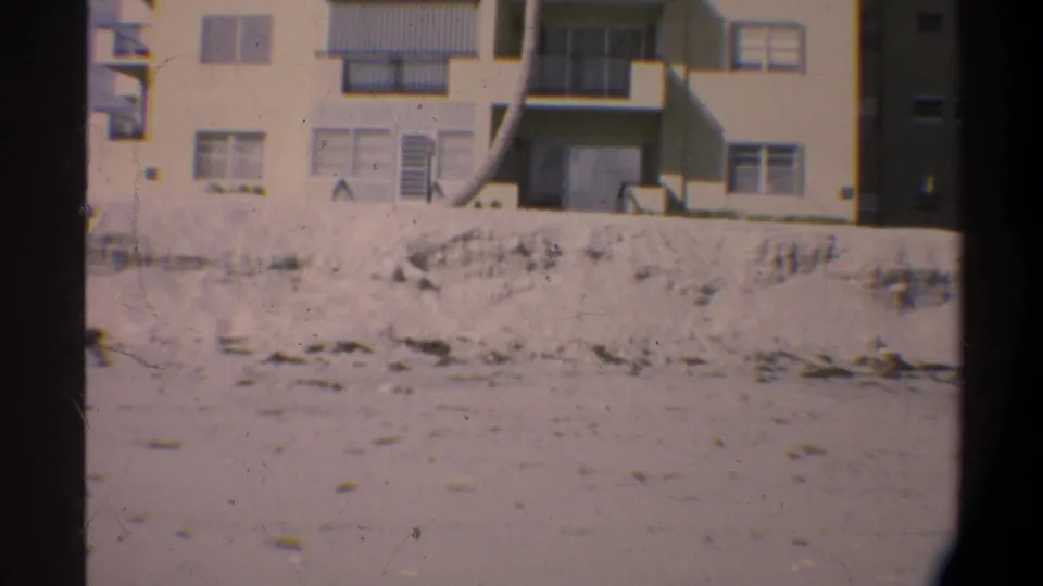 Camera pan reveals a light-colored multi-story apartment block with grid windows and balconies above a sandy lot, circa 1974 Florida, on 8mm film.