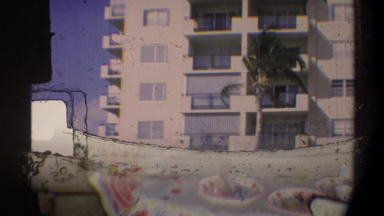 Light-colored multi-rise apartment block with open balconies and a palm tree, circa 1974 Florida; birthday cake partially visible in lower foreground on 8mm film.