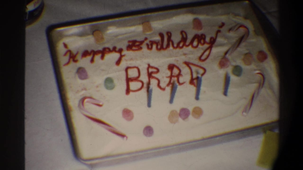 Handmade birthday cake with white frosting, red icing reading 'Happy Birthday BRAD,' candy-cane decorations, multicolored candy dots, and blue candles, circa 1974.
