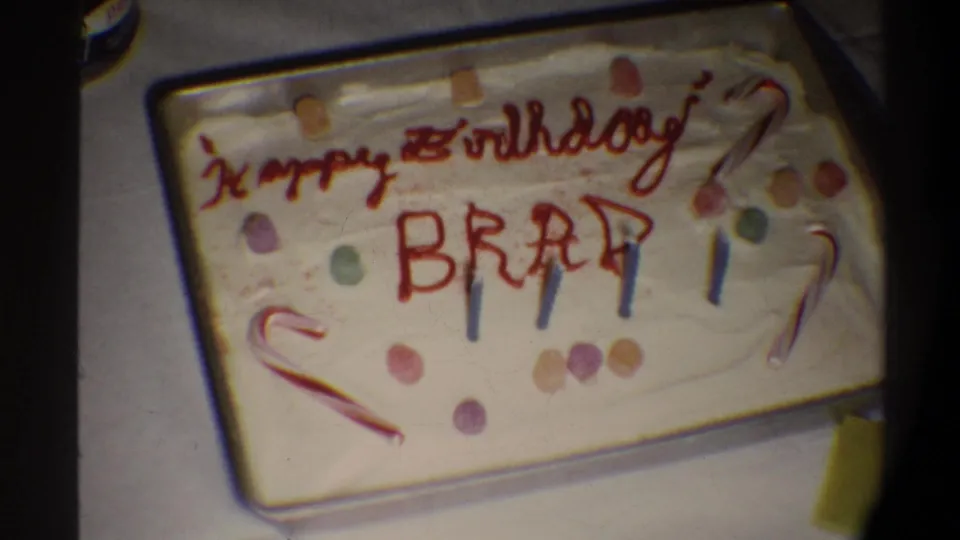 Handmade birthday cake with white frosting, red icing reading 'Happy Birthday BRAD,' candy-cane decorations, multicolored candy dots, and blue candles, circa 1974.