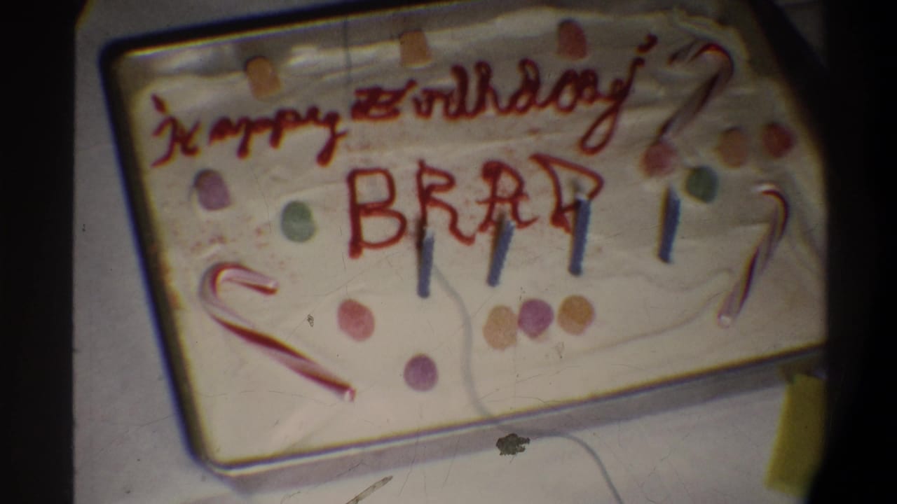 8mm home-movie frame circa 1974, Gainesville FL — sheet cake in a metal pan inscribed 'Happy Birthday BRAD' in red icing, with blue candles, a candy cane, and gumdrops.