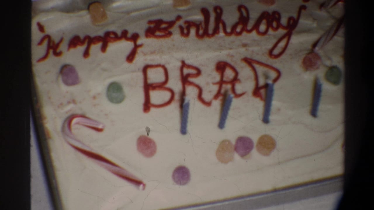 8mm home-movie close-up circa 1974, Gainesville FL — cake surface with red-icing inscription 'Happy Birthday BRAD,' blue candles, candy cane, and gumdrop decorations.