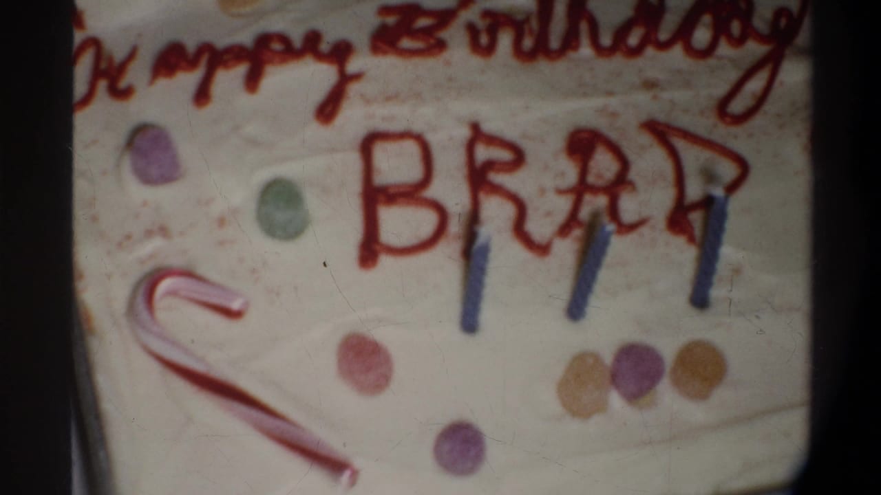 8mm frame circa 1974, Gainesville FL — cake surface with red-script 'Happy Birthday' above block-letter 'BRAD,' blue candles, candy cane, and multicolored gumdrop scatter.