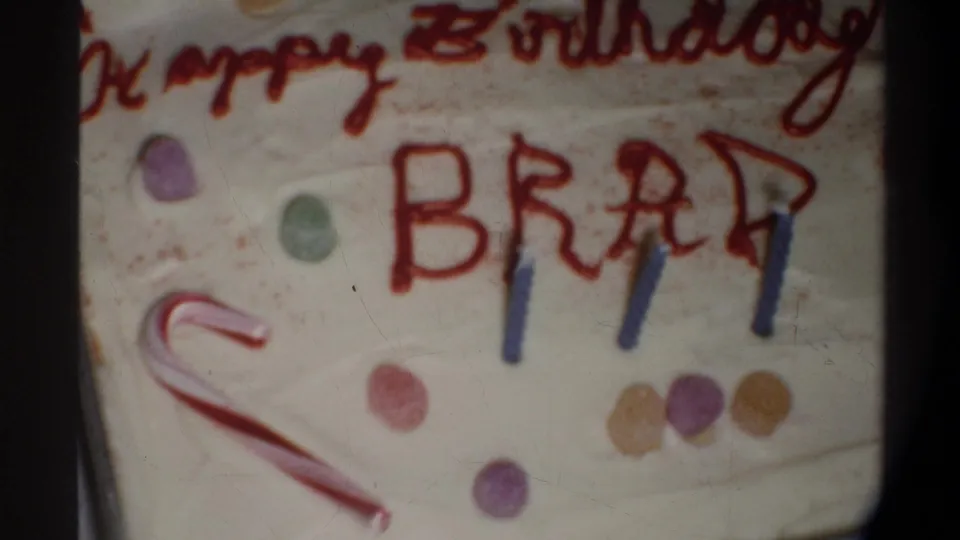 8mm frame circa 1974, Gainesville FL — cake surface with red-script 'Happy Birthday' above block-letter 'BRAD,' blue candles, candy cane, and multicolored gumdrop scatter.
