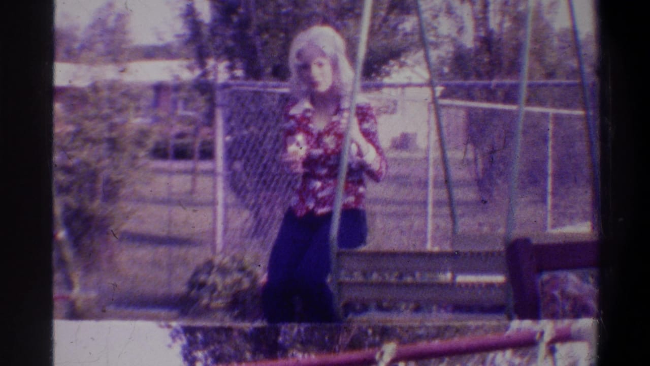 Blonde woman in floral top standing near swing set, chain-link fence and trees behind her, slightly different pose from first frame.
